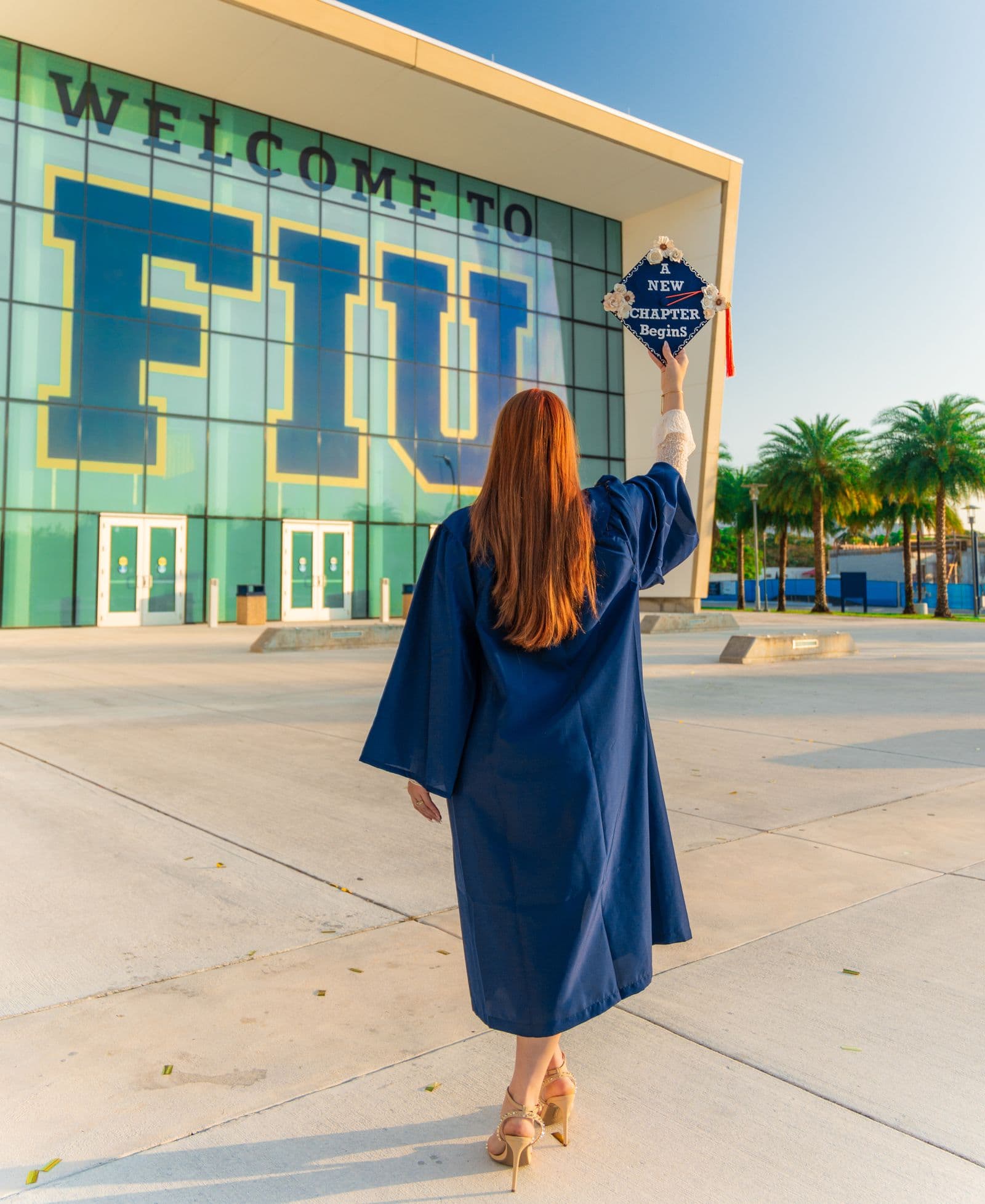 An FIU graduate raises her decorated cap in front of the Welcome to FIU building at golden hour